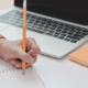 Close up of a hand holding a pencil writing in a notebook in front of a laptop