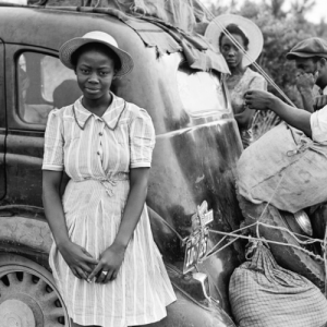 Vintage black and white photograph. A black woman in a dress and hat is standing in front of an old car