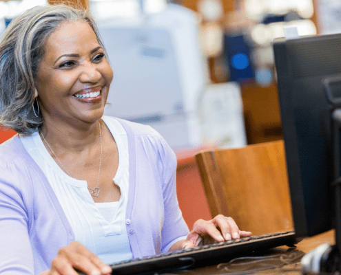 Older Black woman smiling as she uses a desktop computer in the library.