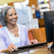 Older Black woman smiling as she uses a desktop computer in the library.